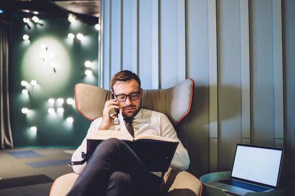 Busy serious businessman talking with client via mobile phone checking information in notebook, male proud financier having cellphone conversation using textbook near table with laptop with mock up