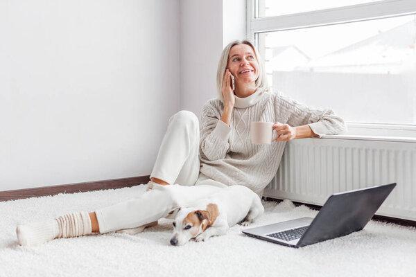Happy middle aged woman with dog using her laptop and mobile phone at cozy white home. Female freelancer working in living roo