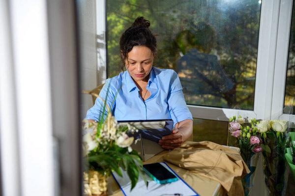 Charming female florist, event manager, flower designer sketching, drawing floral compositions on digital tablet, working in a flower shop. Arrangements and gifts. Creative people and small business