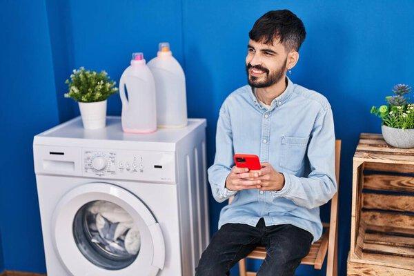 Young hispanic man using smartphone waiting for washing machine at laundry room