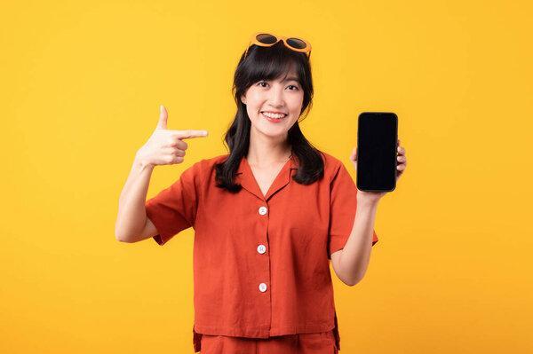 Portrait young beautiful asian woman happy smile dressed in orange clothes pointing finger hand gesture to smartphone isolated on yellow studio background. app smartphone concept