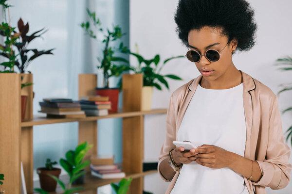 attractive african american businesswoman in sunglasses using smartphone in office