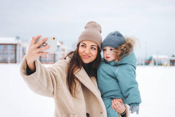 Young pretty mom brunette woman with cute baby boy in winter suit making selfie on street
