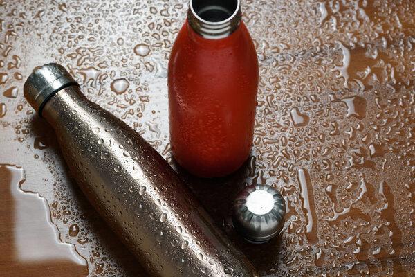 Two stainless thermos bottle on a wooden table sprayed with water