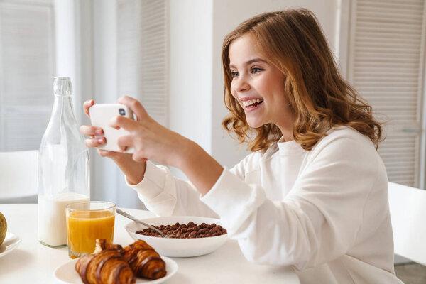 Photo of happy nice girl taking photo on mobile phone while having breakfast in modern kitchen