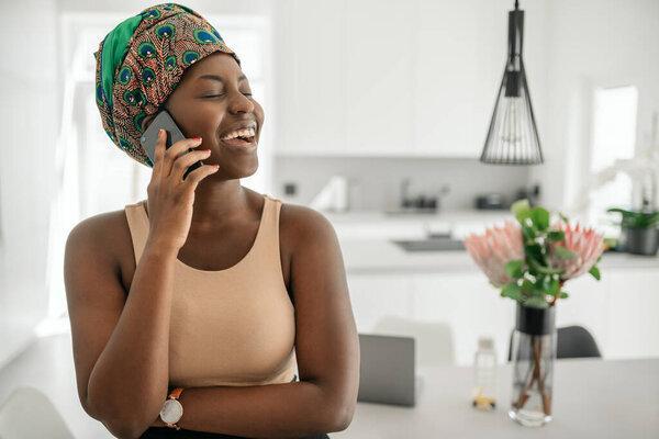 Beautiful African woman at home, smiling and laughing, using mobile smart phone, wearing traditional headscarf and looking into distance with copy space