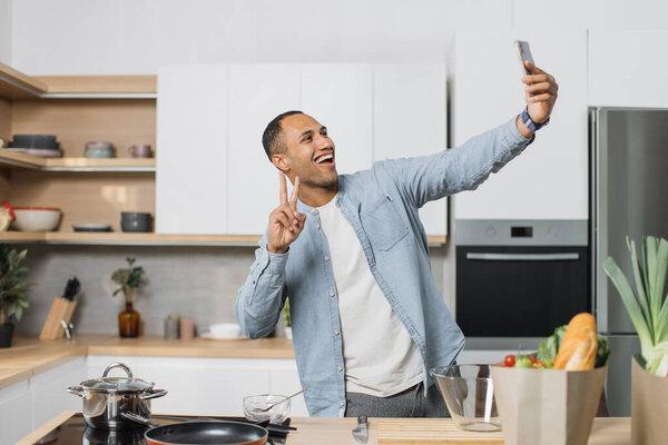 Happy young arabian man preparing dinner searching vegetable recipes diet menu cookbook app using smartphone. Smiling male doing selfie or having video call while cooking healthy vegan food