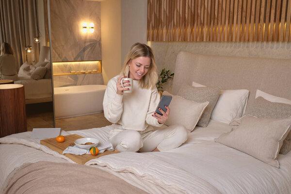 Smiling woman in white pajama sitting on bed with legs crossed and surfing mobile phone while eating breakfast in bedroom at home on weekend