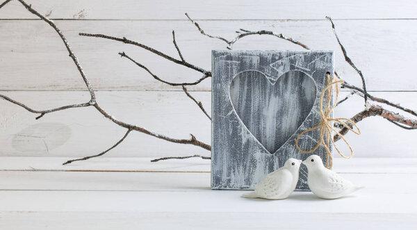Home interior decor in honor of Valentines Day with porcelain figurines of white kissing pigeons birds on background of branches covered with hoarfrost and shabby chic photo frame with heart