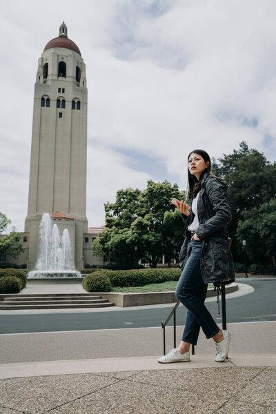 college girl sitting on top of stairs handrail waiting for her boyfriend after course. female asian student using cellphone standing outdoor stanford. church with fountain in background on cloudy day