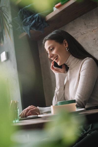 selective focus of green plants and happy freelancer using laptop and talking on smartphone in coffee shop
