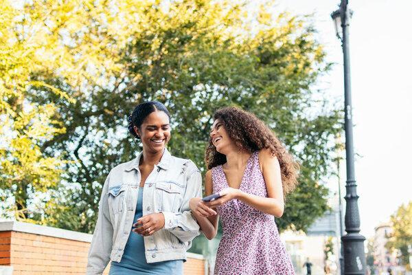 Two Latin American friends walking down the street, having fun, and laughing while using a smartphone.