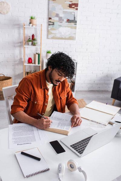 Indian copywriter working with book near devices and papers at home 