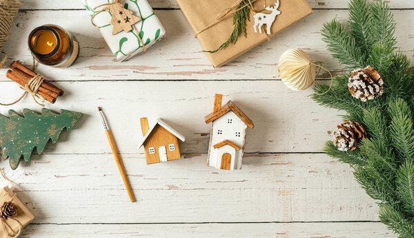 Flat lay of white wooden background with Christmas natural eco decoration. Homemade wooden house with painting brush. Top view gifts, candle, fir tree, toy, cinnamon, pine cone, paper with copy space