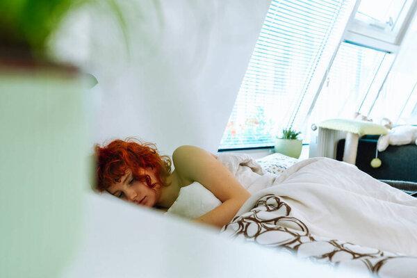 young red-haired woman lies in bed under blanket, uses smartphone, communicates via video conference, reads message, morning vacation concept.