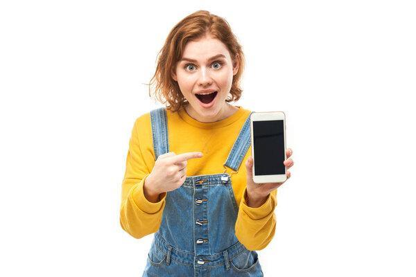 Shocked joyful redhead girl points finger at smartphone blank screen in yellow clothes isolated on white studio background
