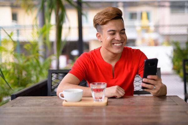 Happy young Asian man using phone at the coffee shop outdoors