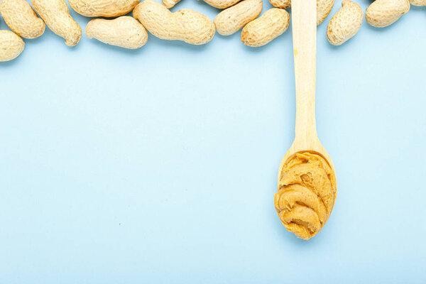 Frame made of nuts peanuts in shell and wooden spoon with creamy peanut butter with copy space on blue colored background. Flat lay with place for text for peanut butter, peanut paste. Vegan food concept