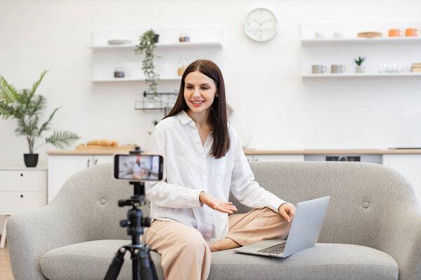 Caucasian woman with brunette hair reading information from modern laptop during live broadcast on social network. Demanding tutor using modern smartphone with tripod for online training.