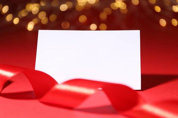 Blank card and ribbon on red background with blurred lights, closeup. Bokeh effect