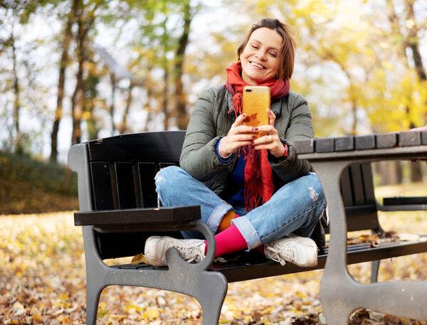 Young adult happy woman 40 years old in bright clothes sits in an autumn park with a smartphone in her hands