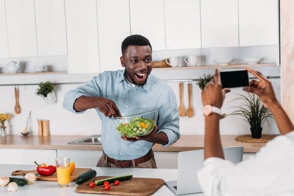 Cropped image of woman taking photo of boyfriend holding bowl with salad
