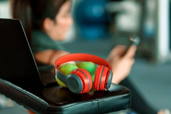 Focus headphone on dumbbell bench and A girl using smartphone in fitness gym background