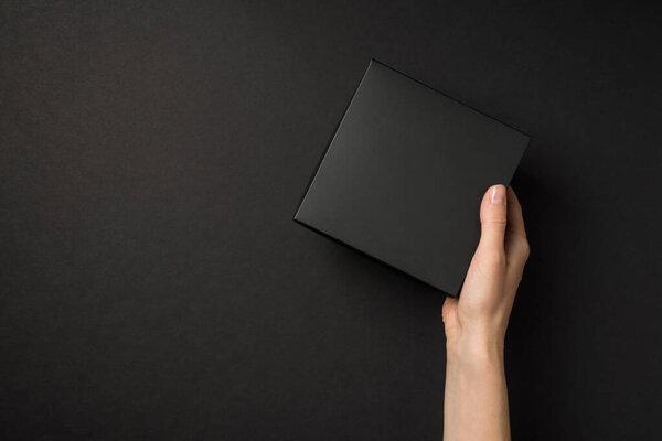 First person top view photo of hand holding black unwrapped giftbox on isolated black background with empty space