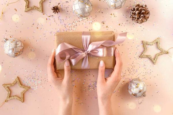Female hands with natural manicure holding a present in kraft paper with pink ribbon. Xmas composition. Flat lay