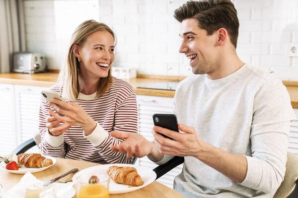 Portrait of smiling couple using smartphones and talking while having breakfast in cozy kitchen at home