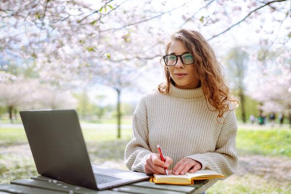 美丽的女人坐在盛开的公园里，用笔记本电脑。Freelance, technology, education, business, blog concept.