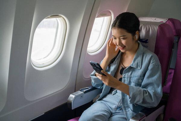 亚洲旅行者女性在飞机上商务旅行期间，坐在窗户附近，一边通过智能手机浏览社交媒体、戴着蓝牙耳机看电影，一边享受着度假式的生活方式。