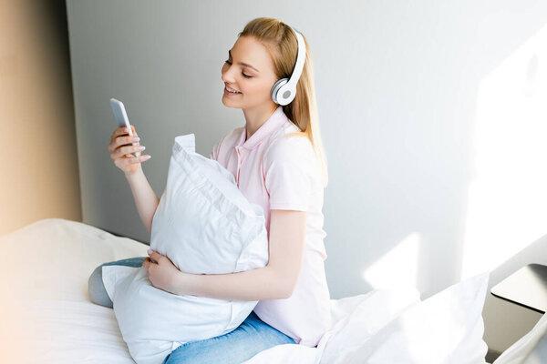 pretty and smiling woman in wireless headphones using smartphone in bedroom