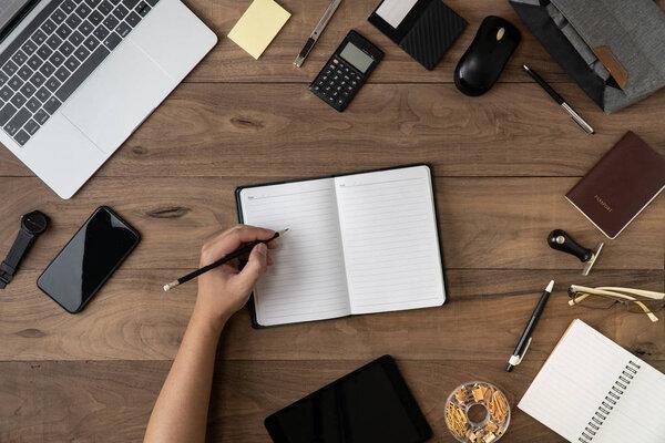 Left hand holding pencil on empty notebook list with office accessories on table flat lay.Working on office desk flat lay.To do list and Meeting reminder list on notebook concept.