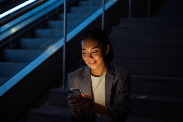 Young smiling asian woman wearing suit using mobile phone while sitting on stairs indoors