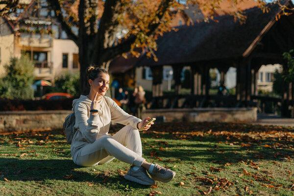 Young fashionable teenage girl with smartphone in park in autumn sitting at smiling. Trendy young woman in fall in park texting. Retouched, vibrant colors. Beautiful blonde teenage girl wearing casual
