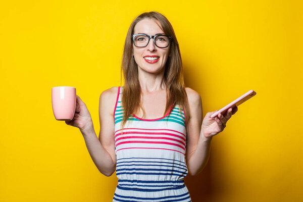 Beautiful young smiling woman holding a phone and a cup with coffee on a yellow background.