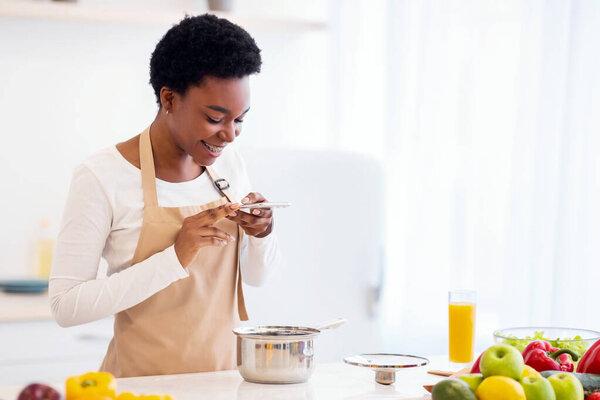 食物博客。非洲女士在平底锅里为准备好的晚餐拍照在家里现代厨房里的电话烹饪。营养博客，家庭休闲概念。体重减轻申请