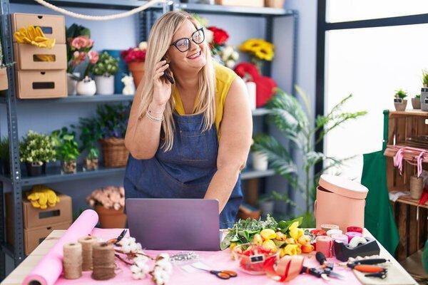 Young woman florist talking on smartphone using laptop at florist