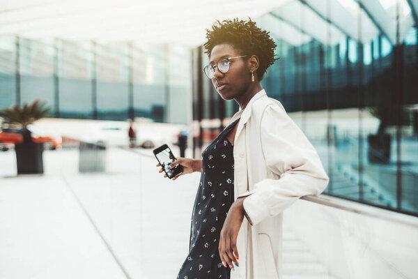 A portrait of a young elegant black hipster female in a white trench and elegant eyeglasses, standing near the entrance of a business center with a smartphone in her hand with its camera turned on