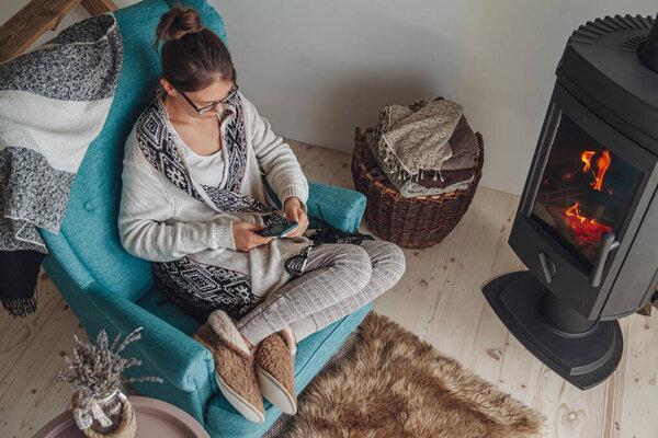 Young woman sitting in a cozy armchair, with a warm blanket, using a mobile phone