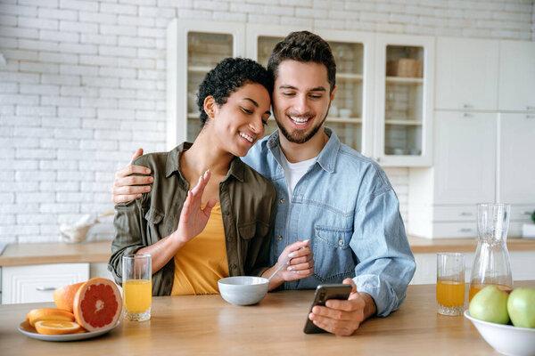 Online video call. Cheerful loving couple of different nationalities, talking via video conference using smart phone, during a morning breakfast, looking into the camera of the mobile phone, smiling