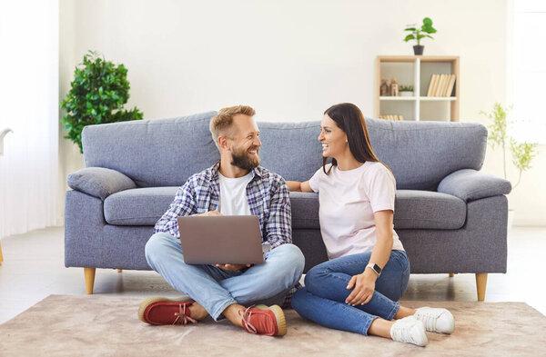 Happy, cheerful family couple using a laptop for online shopping. Smiling married man and woman sitting on the floor by the sofa and choosing home goods in the Internet store on a computer