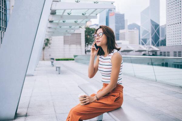 在城市环境中休息的拿着外带咖啡的迷人中国女游客的侧面视图，她正在接听移动电话进行交谈和交流，这是现代科技和 Z 世代的概念。