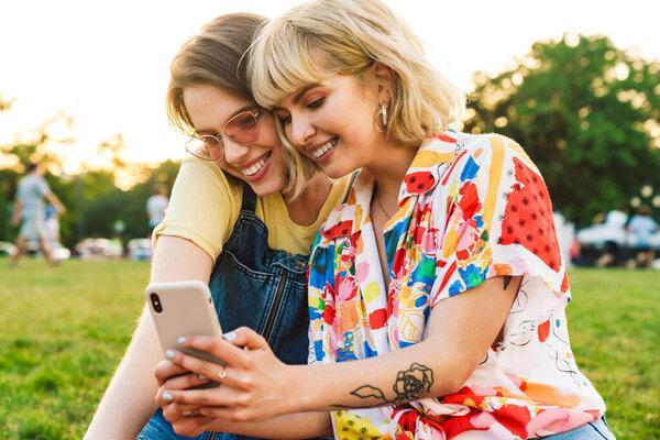 Image of cheerful beautiful two women laughing and sing mobile phone while resting on grass in summer park