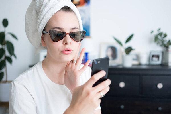 Image of a beautiful woman with towel on head wearing sunglasses chatting and taking photos by mobile phone. Morning routine concept. 