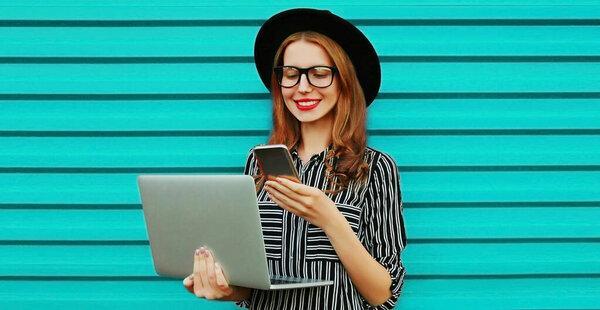 Portrait of happy young woman with laptop looking at smartphone on blue background
