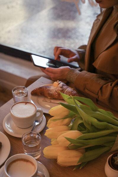 A young girl with a phone in a cafe, on the table is a bouquet of tulips and coffee with buns. Copy space