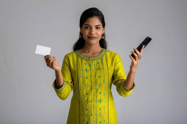 Young smiling beautiful woman or girl presenting credit card while using mobile or smartphone on grey background.