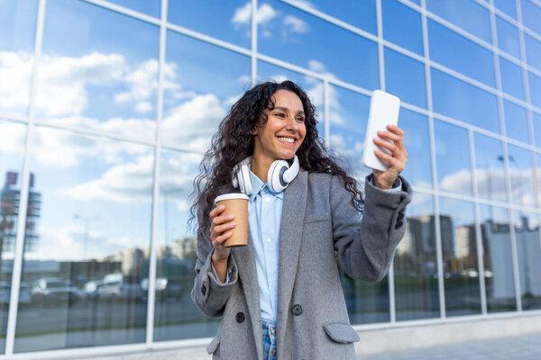 Young beautiful woman walking in the city outside an office building, Hispanic woman in a coat with headphones, a cup of hot drink and a phone, listening to music and browsing the internet.